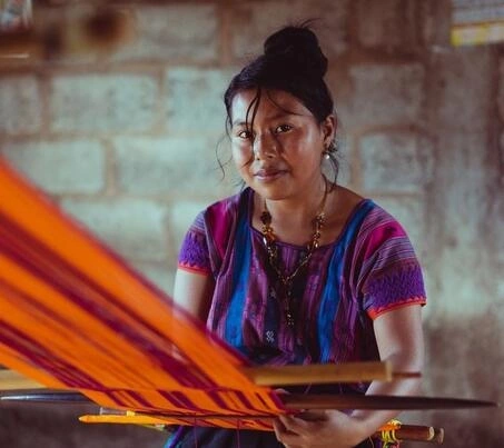 Image of an indigenous woman working on textiles looking at cammera