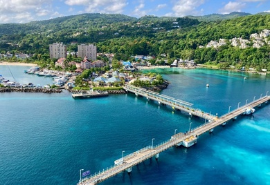 A vibrant coastal scene featuring a turquoise-blue bay with a long pier extending into the water. The shoreline is lined with resorts, marinas, and lush green hills in the background, creating a picturesque tropical setting under a partly cloudy sky.
