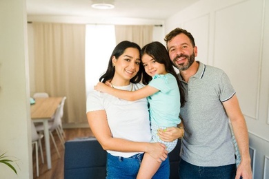 A family standing together in a bright, modern living room with simple furnishings and neutral tones, conveying a comfortable residential setting.