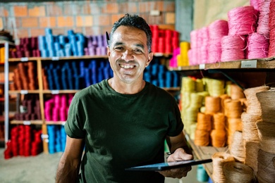 A person stands in a small workshop or warehouse, holding a tablet, with shelves behind them stacked neatly with colorful spools of yarn or thread, suggesting a textile or craft production setting.