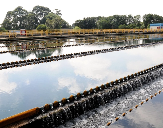 A wide view of a water treatment plant, with multiple parallel concrete channels where water flows evenly over stepped weirs. Yellow safety railings run along the walkways above the channels, and trees frame the background under a clear sky reflected in the water.