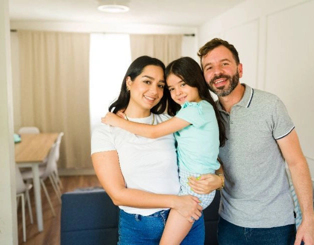 A family standing together in a bright, modern living room with simple furnishings and neutral tones, conveying a comfortable residential setting.