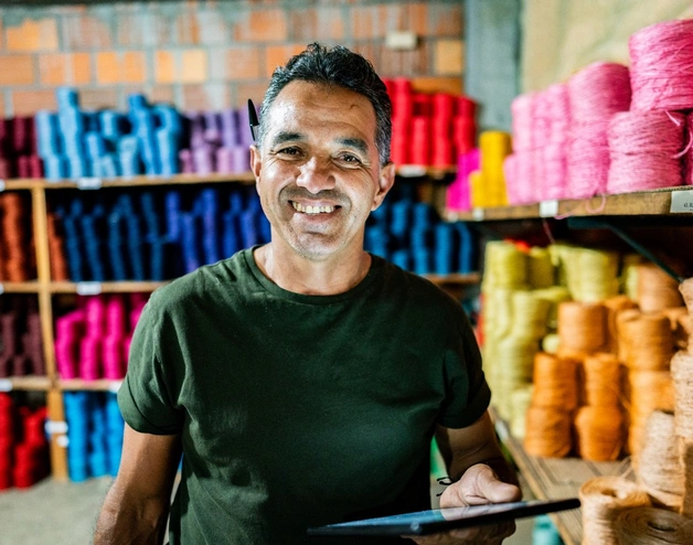 A person stands in a small workshop or warehouse, holding a tablet, with shelves behind them stacked neatly with colorful spools of yarn or thread, suggesting a textile or craft production setting.