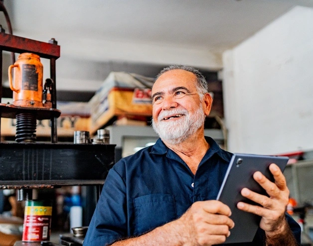 Image of a senior man smiling and holding a tablet while working at his workshop
