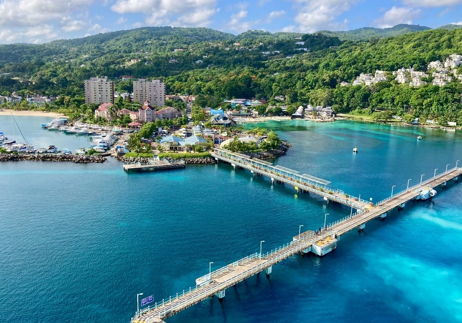A vibrant coastal scene featuring a turquoise-blue bay with a long pier extending into the water. The shoreline is lined with resorts, marinas, and lush green hills in the background, creating a picturesque tropical setting under a partly cloudy sky.