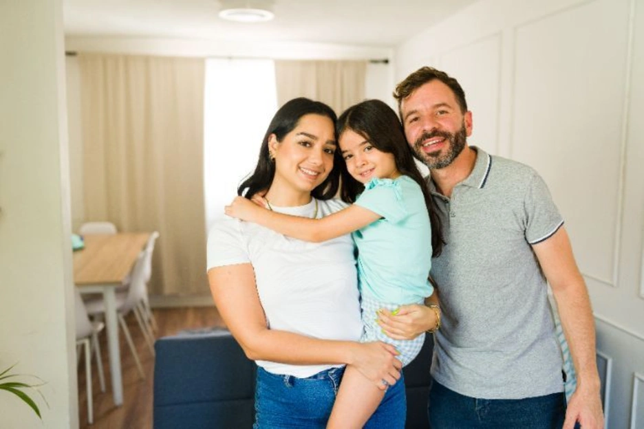 A family standing together in a bright, modern living room with simple furnishings and neutral tones, conveying a comfortable residential setting.