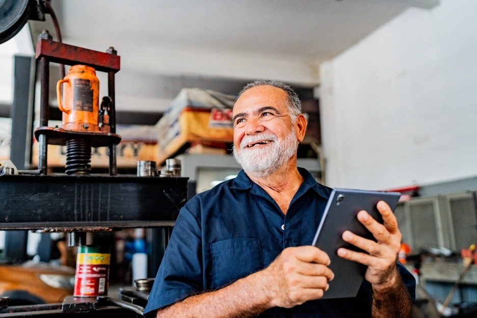 Image of a senior man smiling and holding a tablet while working at his workshop